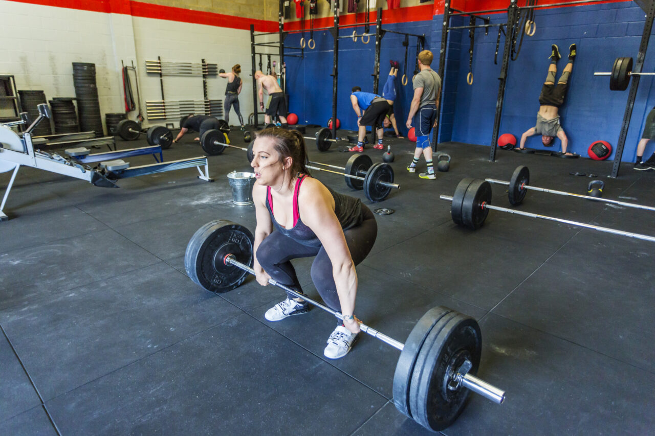Woman lifting barbell in a deadlift at the gym, with other gym goers working our behind her.