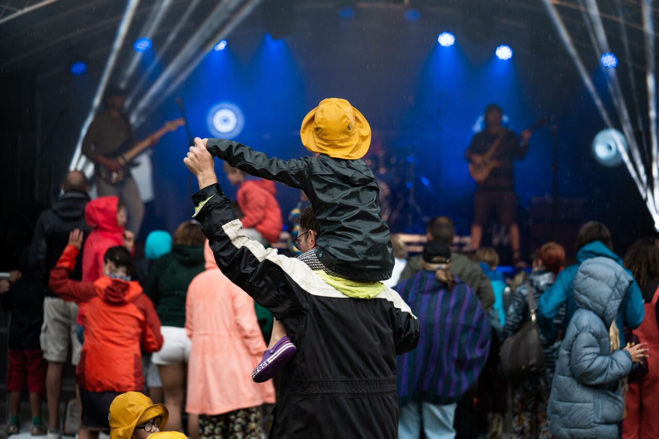 Child sitting on a person's shoulders as they watch a person play guitar on stage.