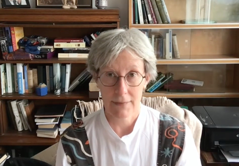 Gwyneth Lewis yn eistedd o blaen silff llyfrau.