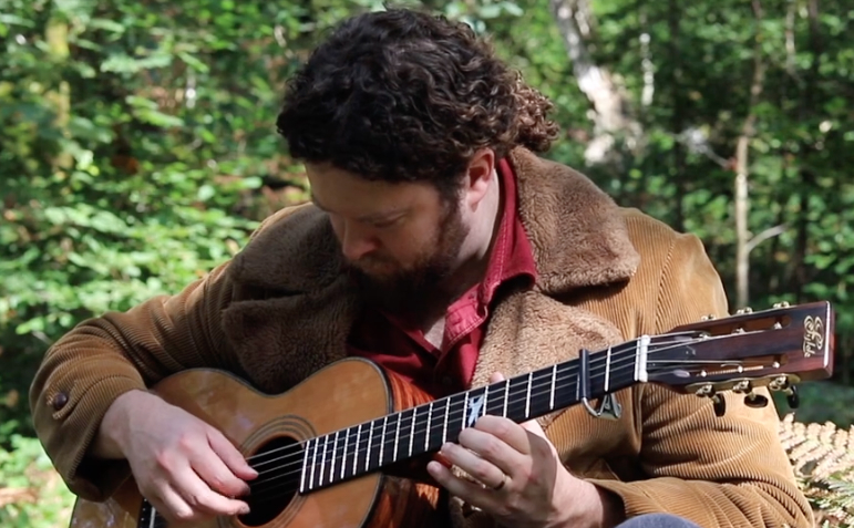 Person yn eistedd tu fas ac yn chwarae gitar.