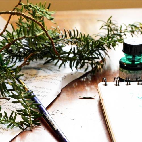 Mae sawl eitem wedi'u gwasgaru ar fwrdd bach: Cangen coeden fechan ar ddarn o bapur crychlyd; potel fach o inc; brwsh paent; pad braslunio. 