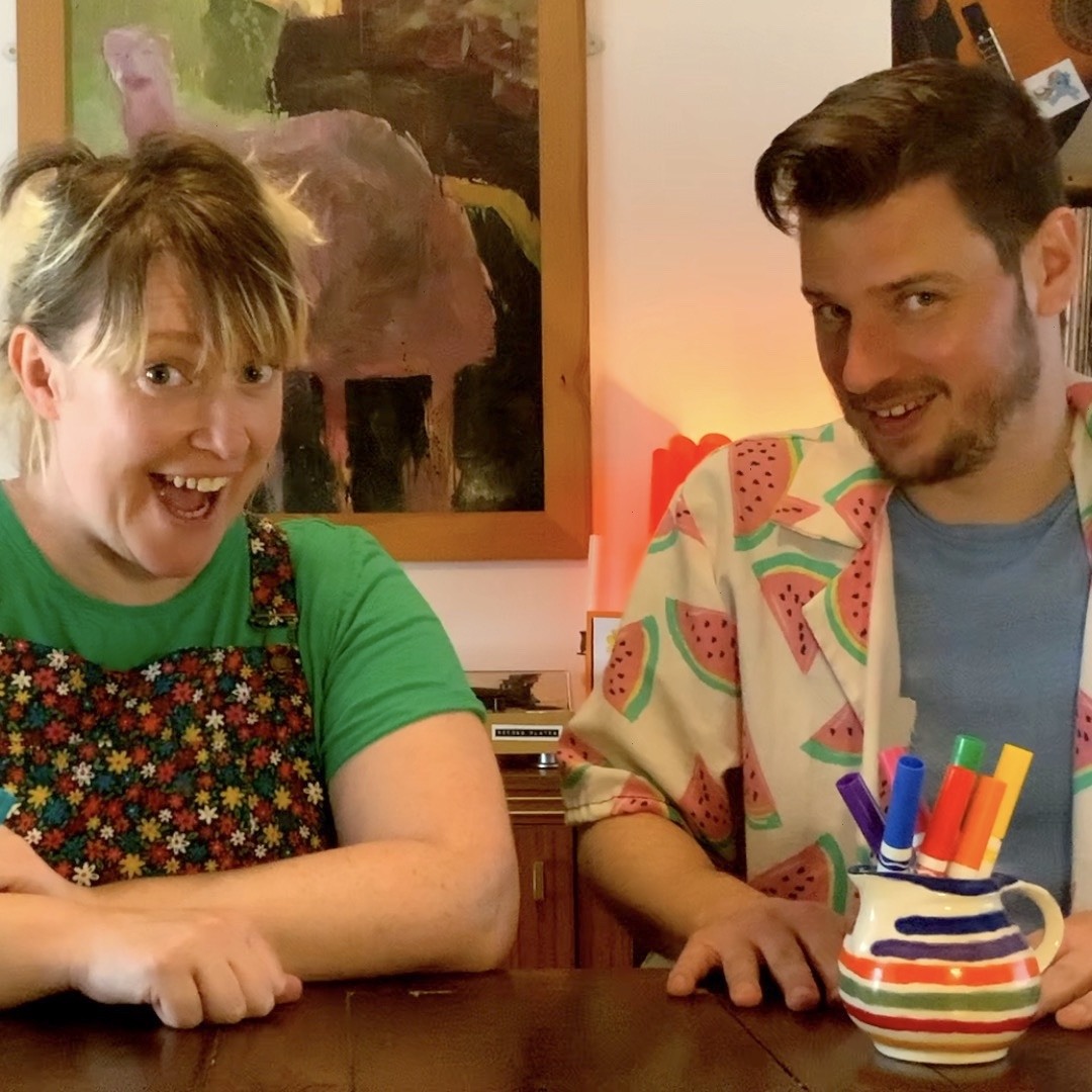 Two people sitting next to each other at a wooden table. One is has blond hair pulled back, the other has short dark hair and a beard. They're both wearing colourful clothing and smiling at the camera.