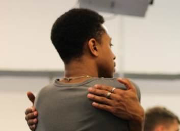 Llun pen ac ysgwydd o berson yn cofleidio'i hun yn dynn. Mae’n nhw'n gwisgo crys llwyd tywyll a modrwyau arian. 