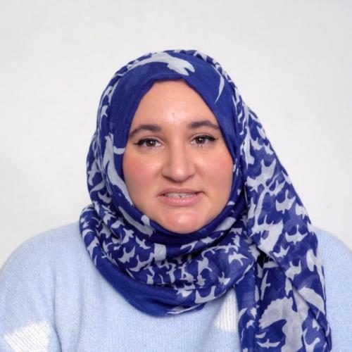 Portrait of a person against a white background wearing a blue and white hijab and a pale blue jumper.