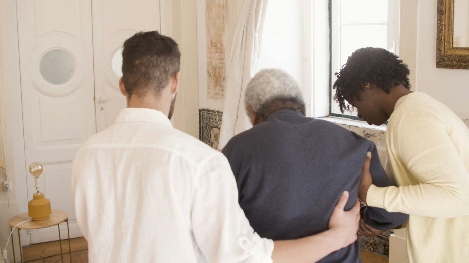 Delwedd o ddyn oedrannus yn cael ei helpu gan ofalwyr